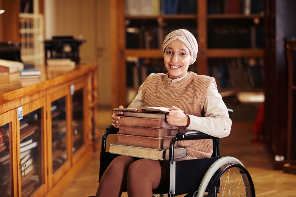 Warm toned portrait of smiling young woman using wheelchair in college library and looking at camera